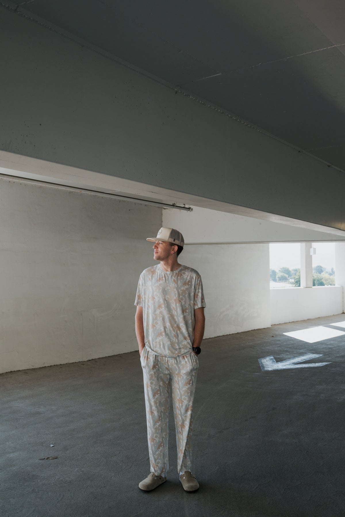 A man wearing the forever french baby Men’s Bamboo Short Sleeve Pajama in the Woodland print, paired with a matching cap and slippers, stands in a parking garage with his hands in his pockets, looking to the side as light streams through windows behind him.
