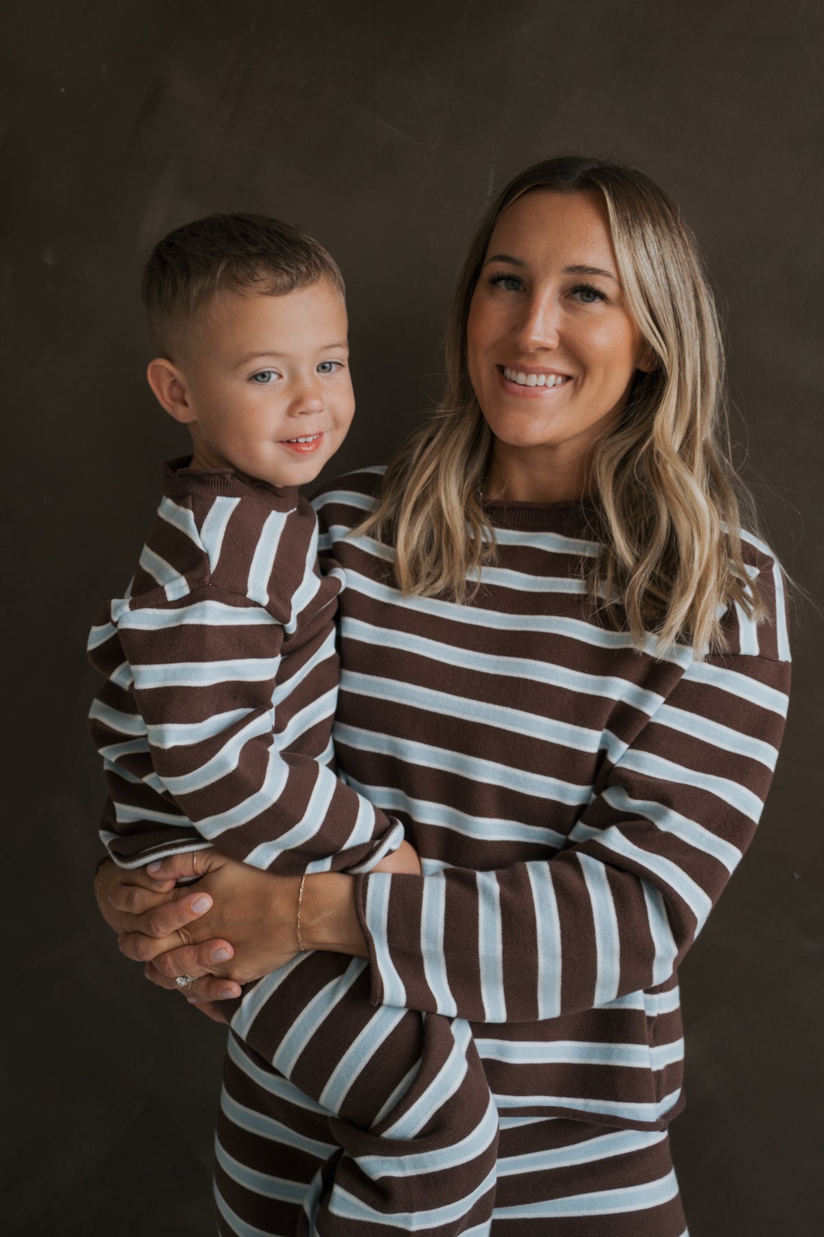 A smiling woman holds a young boy, both wearing matching brown and light blue striped Knit Pant Set | Garrett *PRE-ORDER* long-sleeve knit sets.