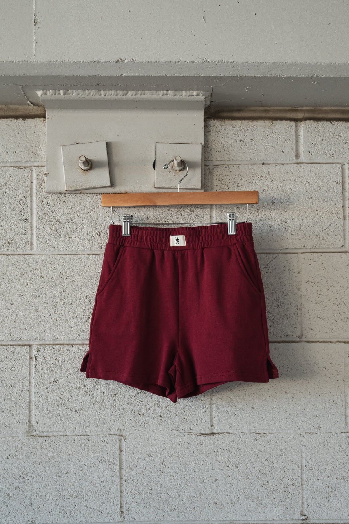 Forever French Baby Women’s Terry Shorts in deep cranberry with an elastic waistband, hanging on a wooden hanger against a white brick wall.