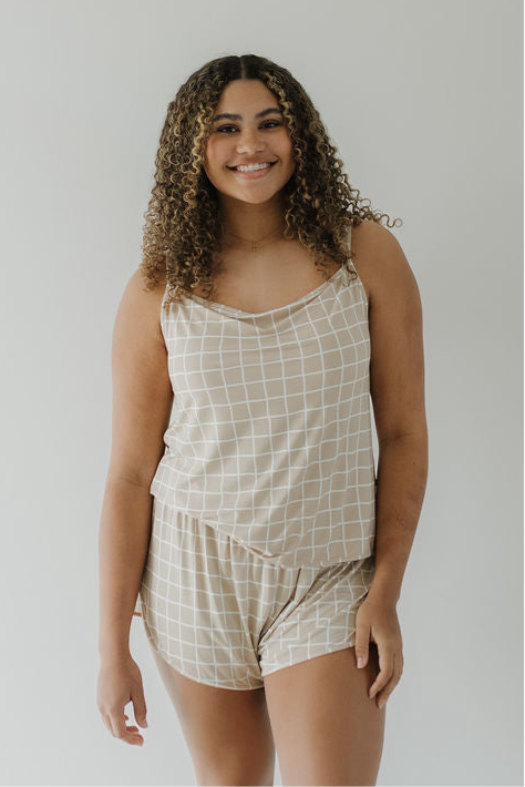 A woman with curly hair smiles in the Cami Women's Bamboo Set | Grid by forever french baby, featuring a breathable beige tank top and shorts with a white grid pattern, against a plain light background.