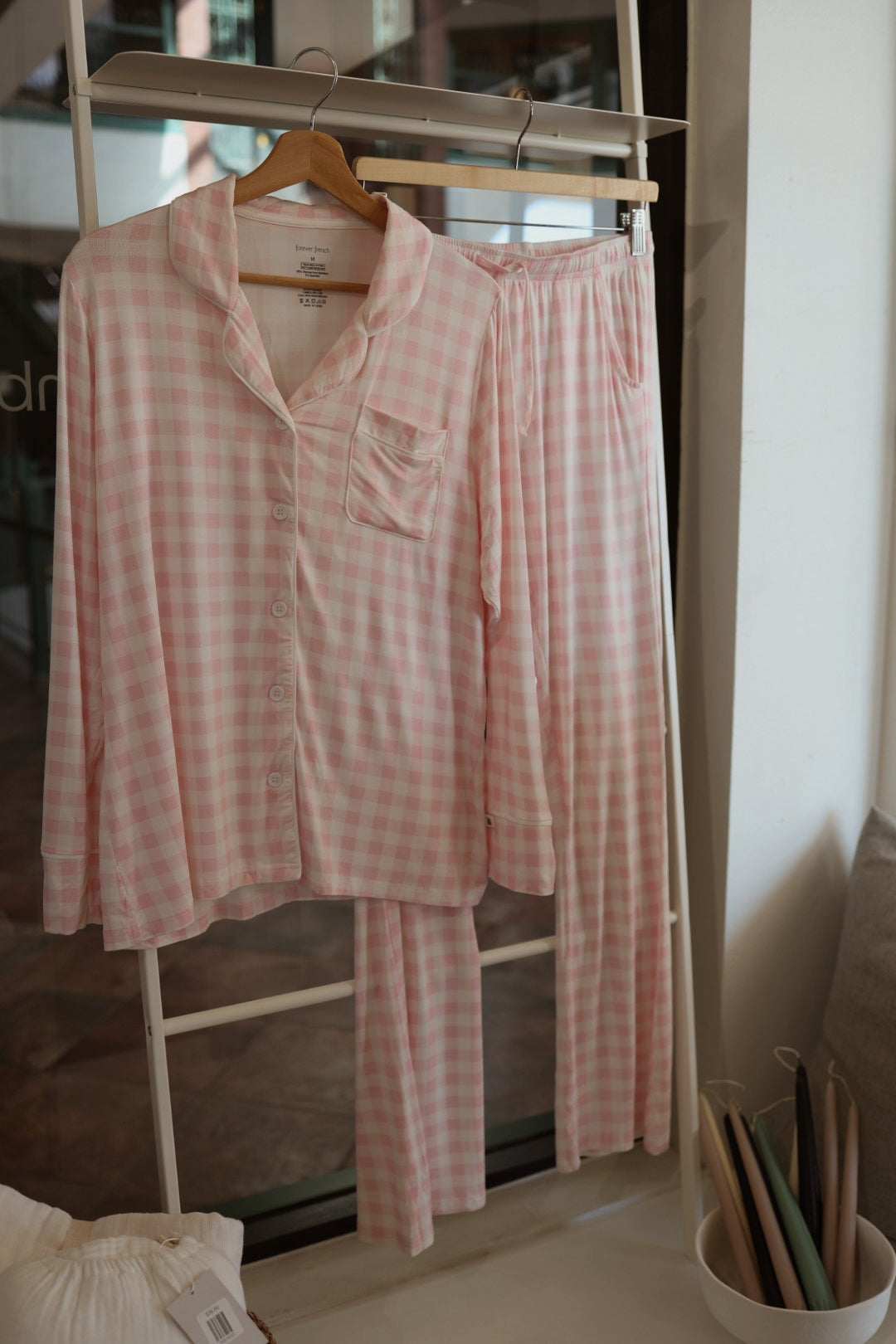 The Long Sleeve Women's Bamboo Pajama in Pink Sugar by forever french baby, featuring a pink and white checkered button-up top and matching pants, hangs on a wooden hanger near a window with pastel folders stacked nearby.