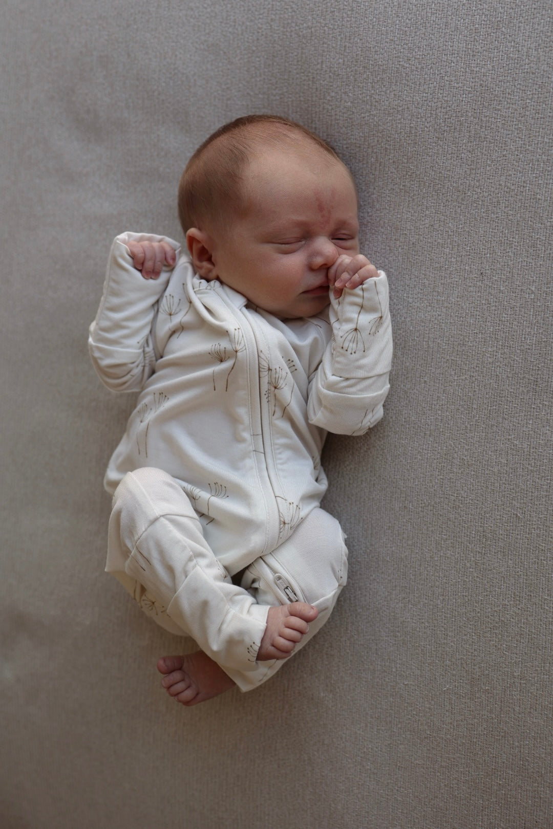 A newborn sleeps peacefully on a soft, light grey surface, wearing forever french baby’s Bamboo Zip Pajamas in the Dandelion print. The white onesie with subtle patterns keeps them cozy, with tiny hands curled by their face and bare feet.