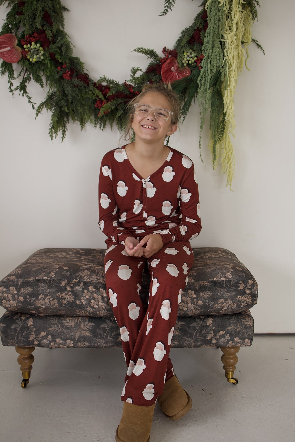 A child wearing red Youth Flare Bamboo Pajamas | Vintage Santa with a white Santa pattern sits on an upholstered bench in front of a greenery garland.