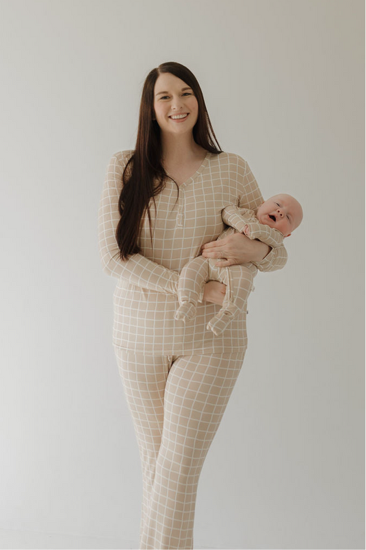 A woman with long brown hair smiles and stands against a plain light background, wearing forever french baby Women's Bamboo Pajama | Grid, while holding a baby in matching checkered pajamas.