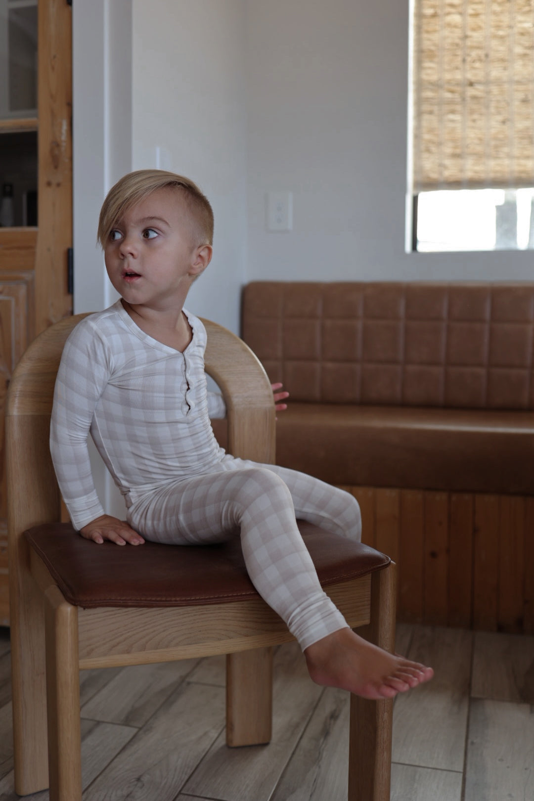 A young blonde child in forever french baby's Bamboo Two Piece Pajamas | Gingham sits sideways on a wooden chair with a brown cushion, gazing left in a cozy room featuring a bench and window.