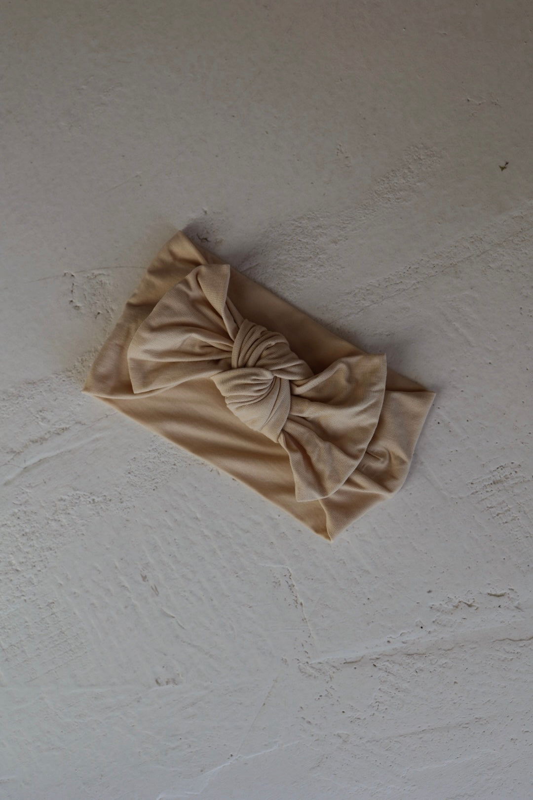 A Bamboo Head Wrap in Bone by forever french baby, featuring a large twisted bow detail, lies flat on a textured, light surface.