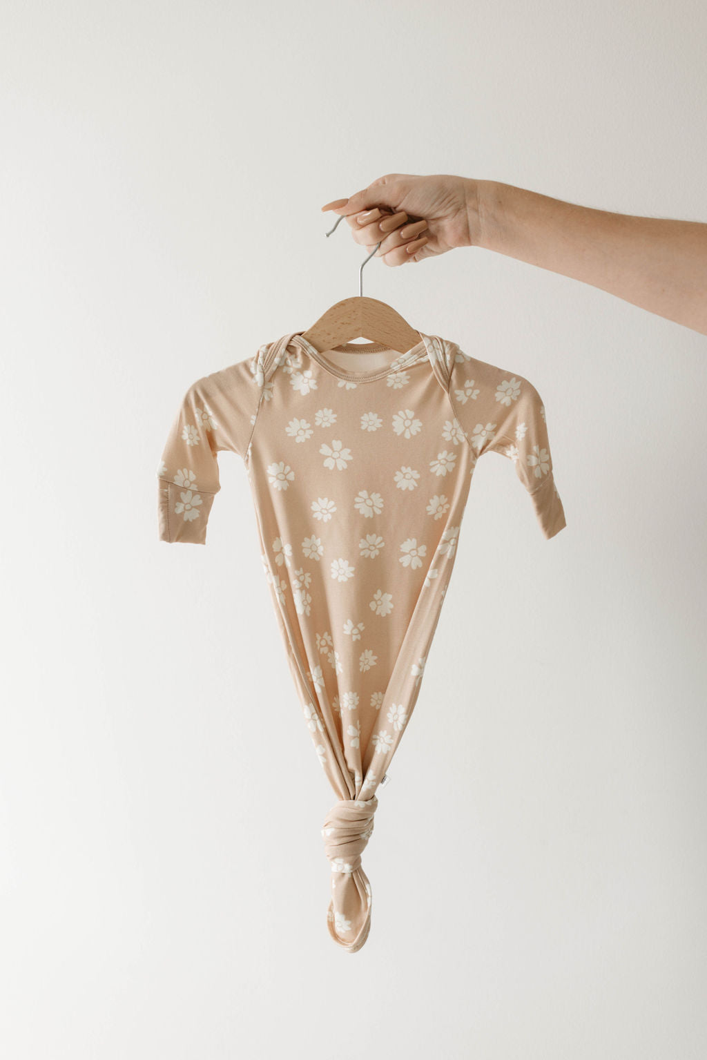 A hand holds the forever french baby Lazy Daisy Bamboo Knotted Gown, featuring a beige color with white daisy patterns and a knotted bottom, set against a plain white background.