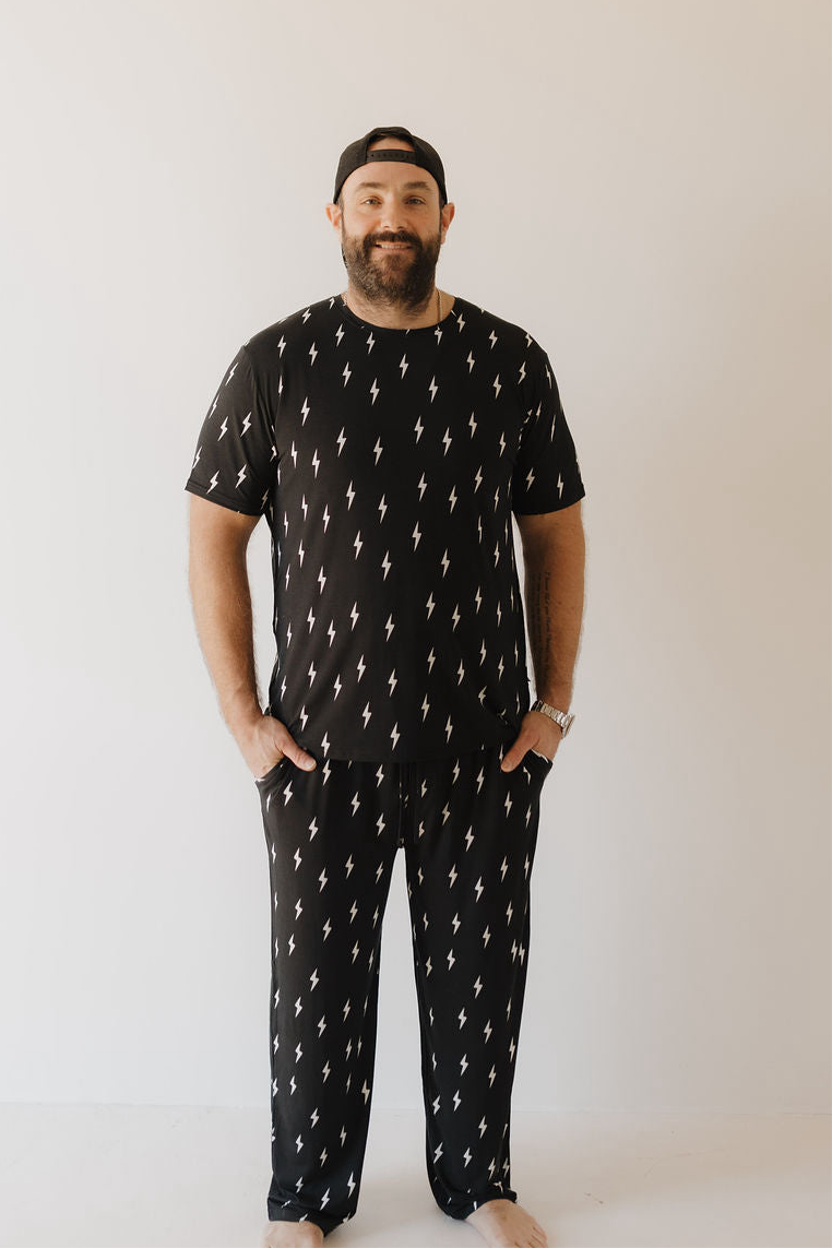 A bearded man smiles barefoot against a white background, hands in pockets, wearing the Men's Bamboo Short Sleeve Pajama in Midnight Lightning Bolt by forever french baby, showing off its breathable comfort and stylish lightning bolt design.