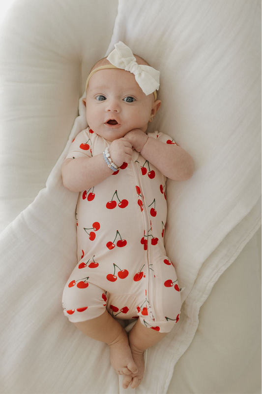 A baby wears the forever french baby Bamboo Zip Shortie | Cherry Orchard, featuring a cream base with red cherry prints and a large white bow headband, lying on a soft white blanket and wrapped in breathable bamboo fabric.