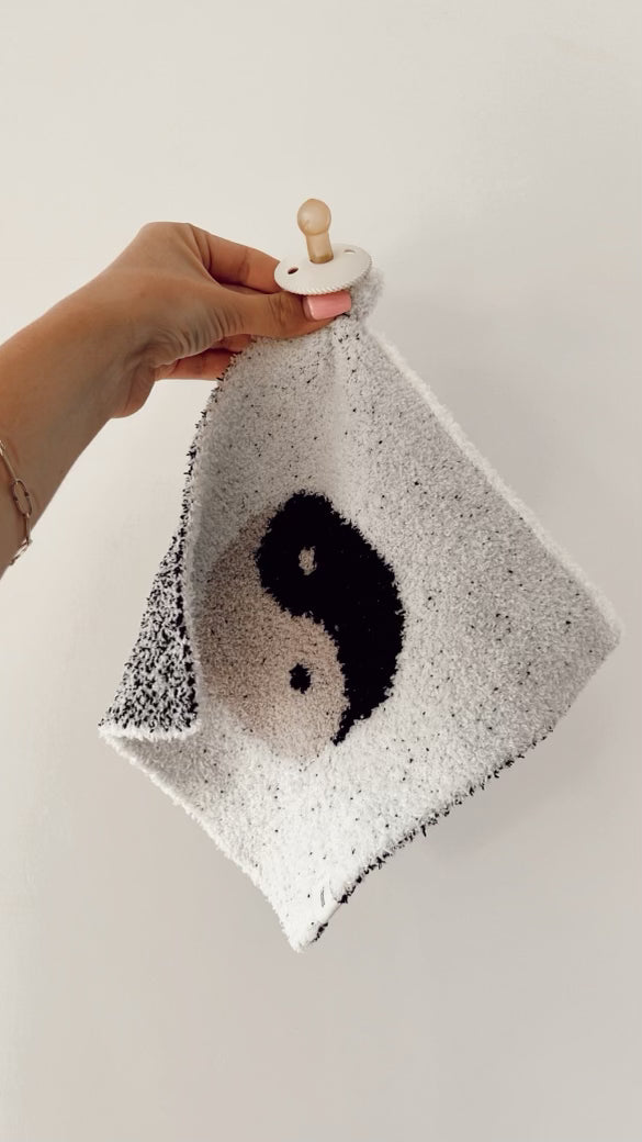 A hand is holding the forever french baby Lovey | Yin-Yang Black & White, a small square blanket with fuzzy edges made of microfiber feather yarn. The blanket features a black-and-white yin-yang symbol in the center and has an attached white pacifier. The background is a plain, off-white wall.