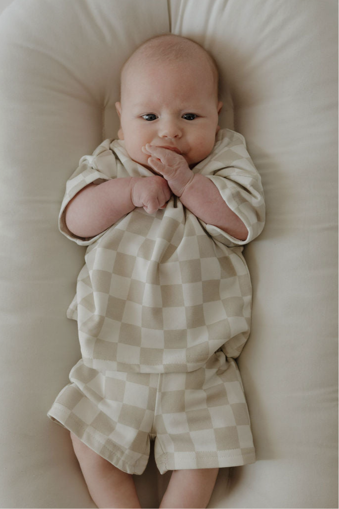 A baby relaxes on a cushioned surface, wearing the forever french baby French Terry Short Set | Checkerboard in beige and white. With one hand near their mouth, the baby looks calm and content.