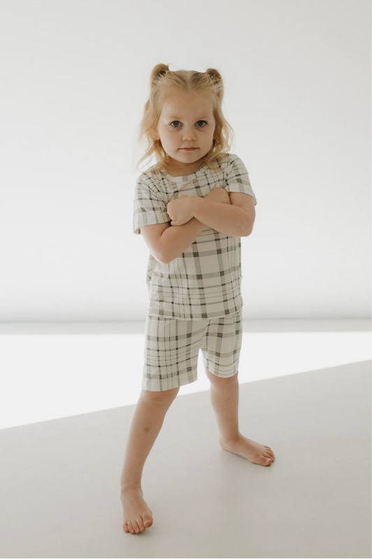 A young girl with blonde pigtails stands barefoot on a white floor, confidently wearing the forever french baby Bamboo Short Set | Picnic Plaid made from breathable sleepwear fabric, arms crossed in front of her chest.