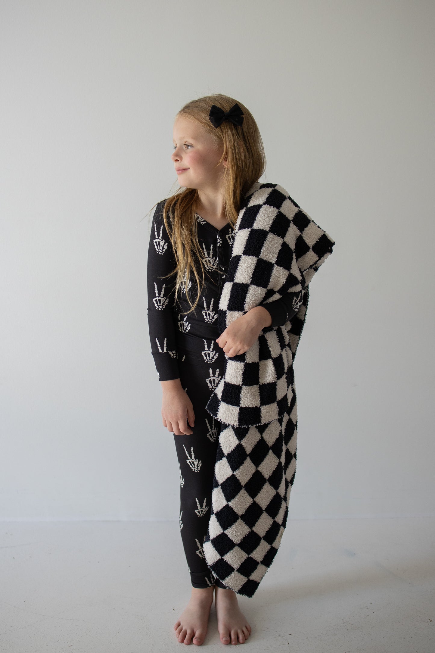 A young girl with long blonde hair stands barefoot against a plain white wall, smiling and looking to her left. She’s dressed in forever french baby’s Bamboo Two Piece Pajamas in the Midnight Skelly print—breathable, stylish sleepwear designed for all-night comfort. She holds a black and white checkered blanket draped over one arm.