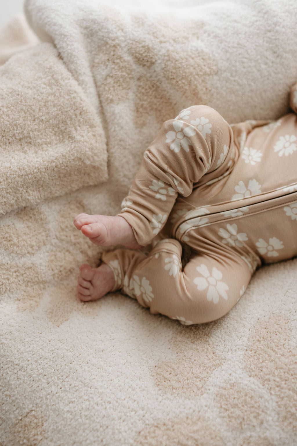 Close-up of a baby's legs and feet, dressed in soft, beige pajamas with a white floral pattern. The baby is lying on the Plush Blanket | Lazy Daisy by forever french baby, showcasing its fuzzy, cream-colored fabric with a velvety-rich texture.