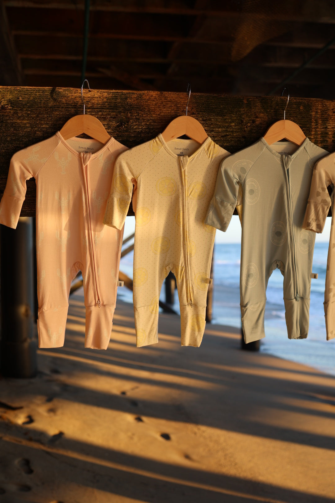 The Bamboo Zip Pajamas | Lobster by forever french baby hang on wooden hangers with sunlight streaming in, showcasing hypoallergenic baby clothing against a sandy beach and ocean backdrop.