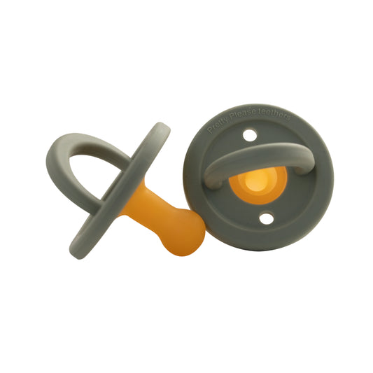 Two baby essentials with a modern design are displayed. One Modern Pacifier | Eucalyptus by forever french baby is positioned upright, while the other is on its side. Both have a grey outer ring and handle with a soft, orange silicone teat. The text "Pretty Please Teethers" is visible on the ring of one pacifier.
