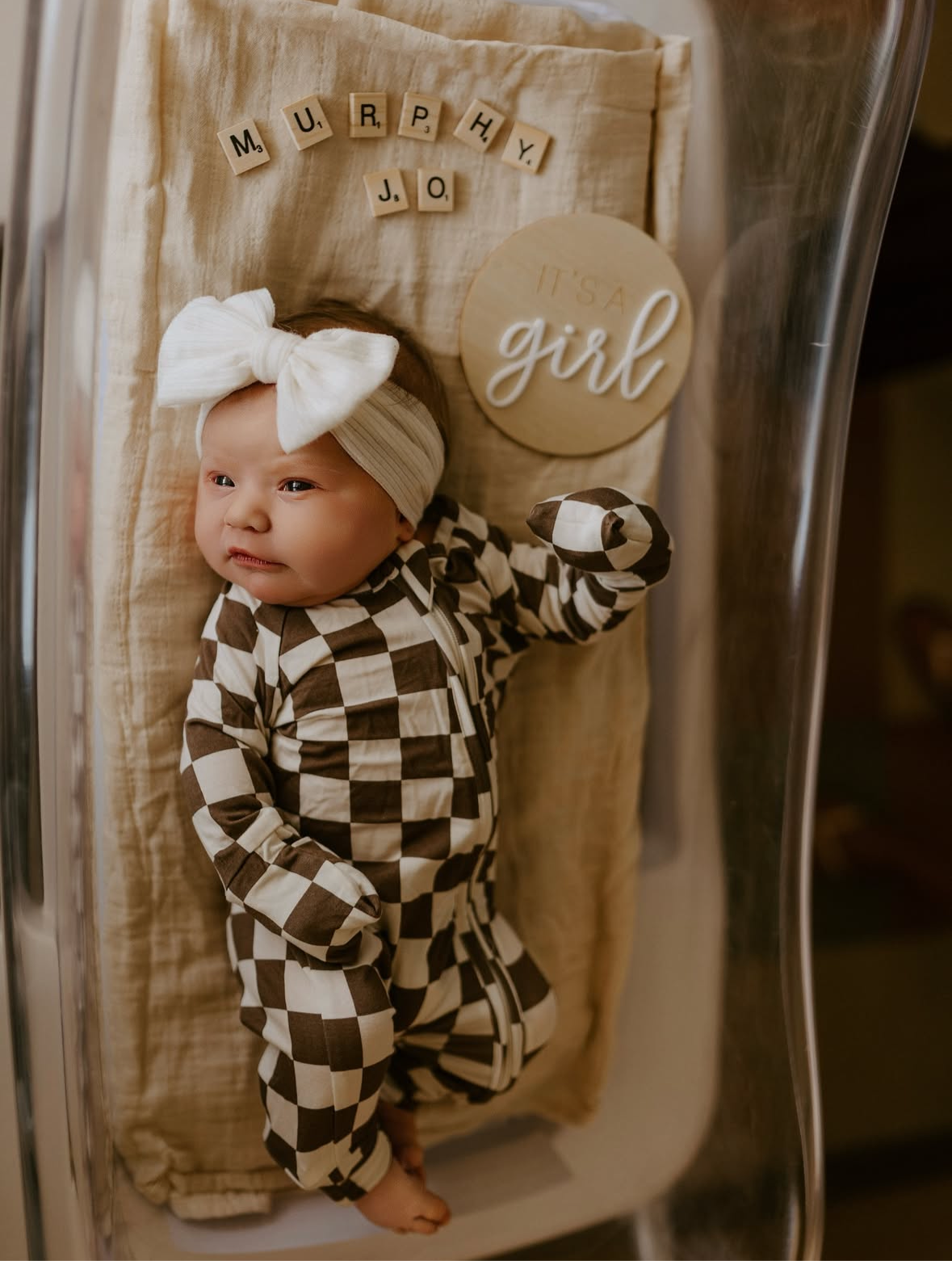 A newborn in a hospital bassinet wears forever french baby Bamboo Zip Pajamas | Dutton—a black and white checkered outfit—and a large white bow headband. Wooden letters spell “Murphy Jo,” with a round sign reading “It’s a girl.”.