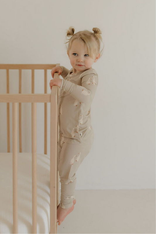 A young child with light hair in two buns stands barefoot by a light wooden crib, wearing forever french baby’s Bamboo Two Piece Pajamas | Silly Goose, featuring a beige bear print, in a minimal white room.