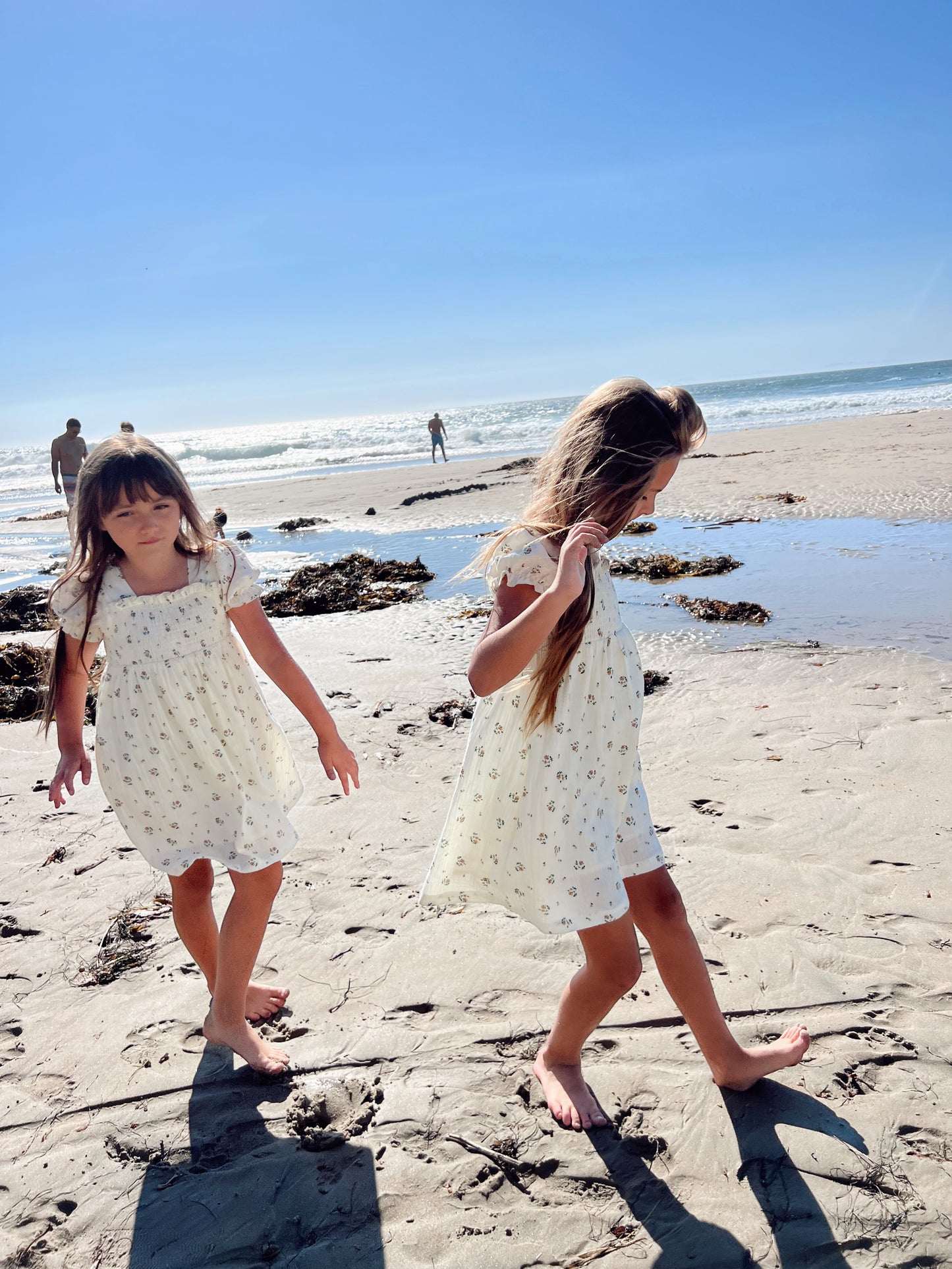 Two young girls with long hair walk barefoot on a sandy beach, their matching ivory Child & Women's Muslin Dresses from the Garden Days collection by forever french baby adorned with delicate patterns. Seaweed is scattered on the sand, and people can be seen in the distance under a clear blue sky.