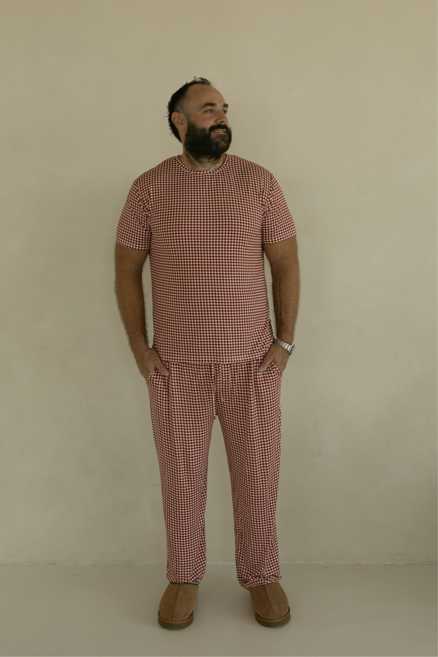 A bearded man stands against a plain light wall, wearing forever french baby's Men's Short Sleeve Bamboo Pajamas in Red Gingham, with hands in his pockets and brown slip-on shoes.