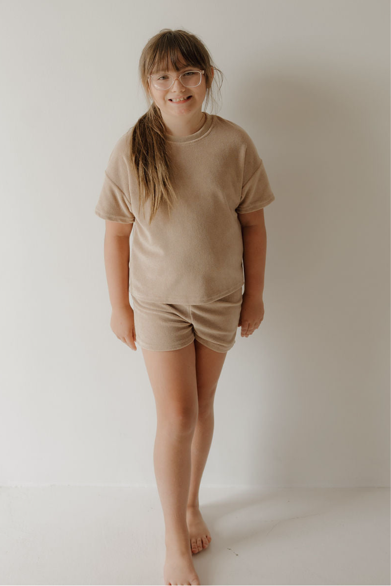 A young girl with long light brown hair and glasses smiles at the camera, barefoot in a forever french baby Child Terry Short Set | Tan. Available in sizes 0 months – 12 years for all your family moments.