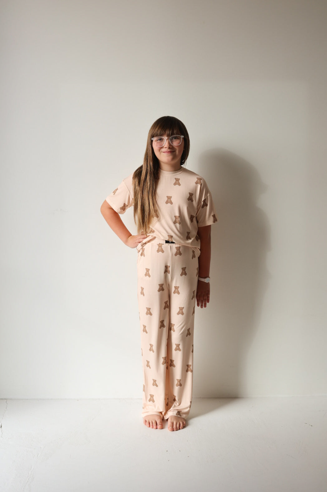 A young girl with long brown hair and glasses stands barefoot against a white wall, smiling in her forever french baby Child Boxy Tee Lounge Set | Lottie—breathable kids sleepwear that keeps her comfy all night.