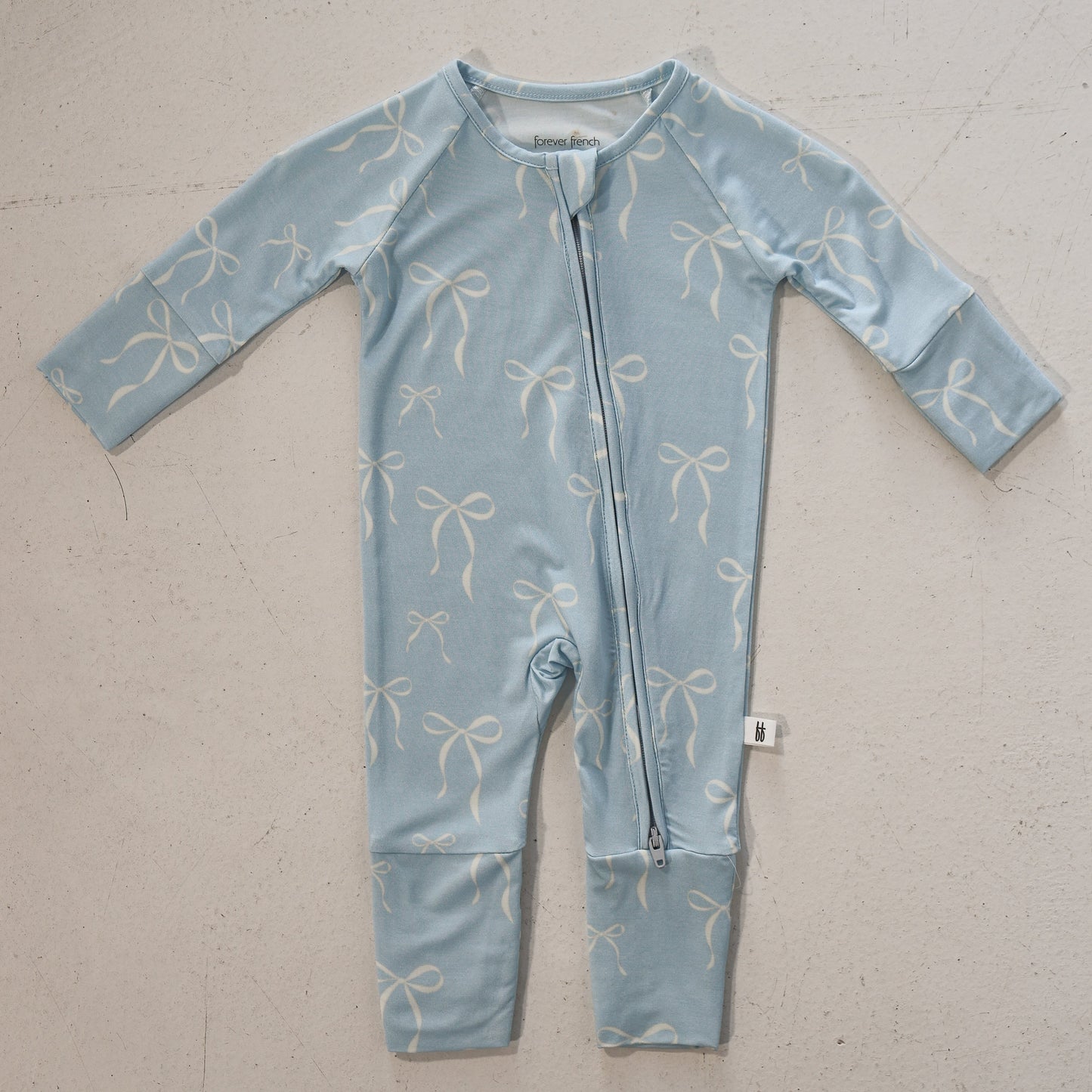 The forever french baby Bamboo Zip Pajamas | Blue Bows is a light blue, hypo-allergenic baby onesie with long sleeves, legs, and a diagonal zipper. Made of breathable bamboo, it features a white bow pattern and is shown laid flat on a light surface.