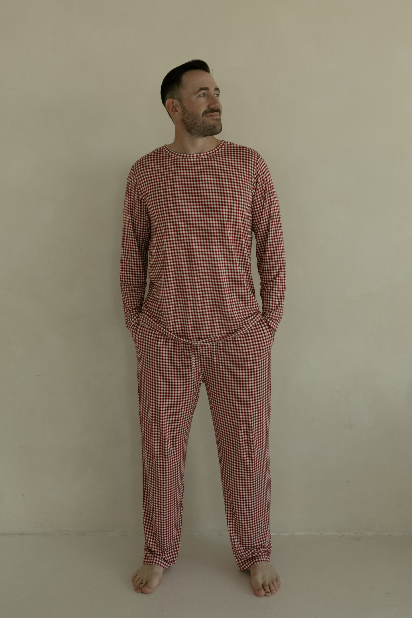 A man stands barefoot by a beige wall, smiling slightly, wearing forever french baby Men's Bamboo Pajamas in Red Gingham with his hands in his pockets.