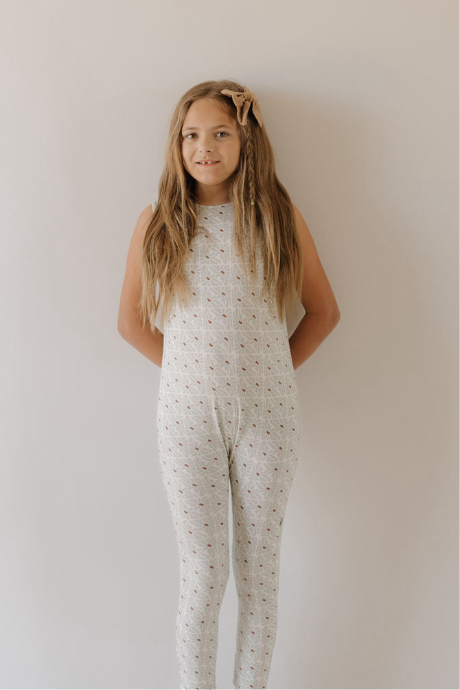 A young girl with long, light brown hair stands against a plain light wall, wearing the forever french baby Harem Bamboo Romper | Coast and a matching bow, her hands behind her back and a slight smile.
