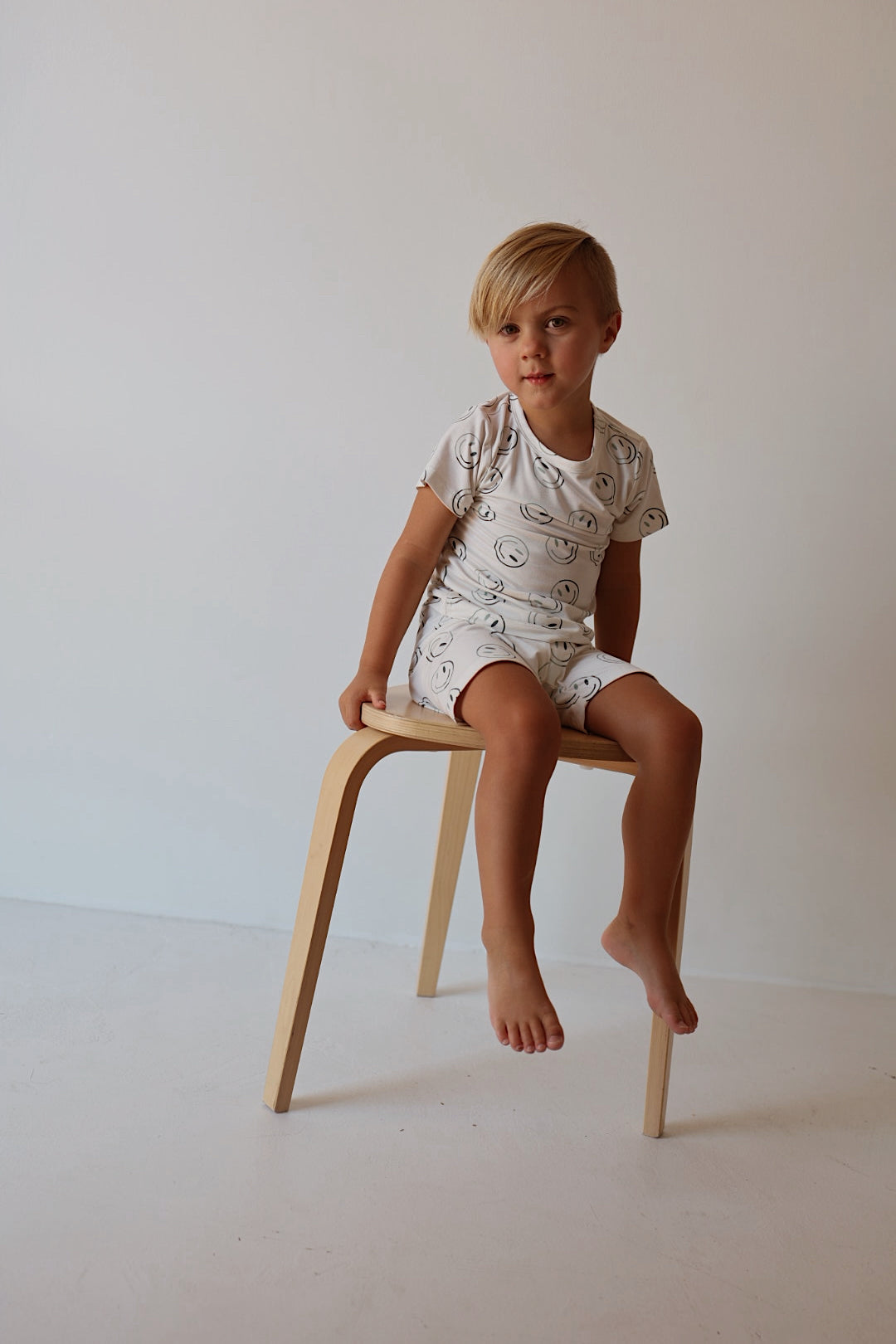 A young blond child, barefoot, wears the forever french baby Bamboo Short Set | Green Smiles—hypoallergenic kids clothing—in white with a simple line pattern, seated on a light wooden stool against a soft, neutral background.