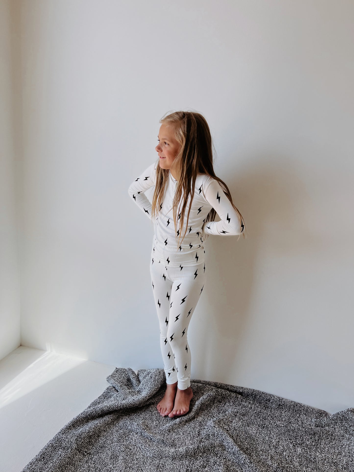 A young child stands on a gray blanket against a white wall, wearing hypo-allergenic Bamboo Two Piece Pajamas | Black & White Lightning Bolt by forever french baby, made from soft bamboo fabric. The child has long, straight hair and stands with hands on hips, looking to the side with a cheerful expression.