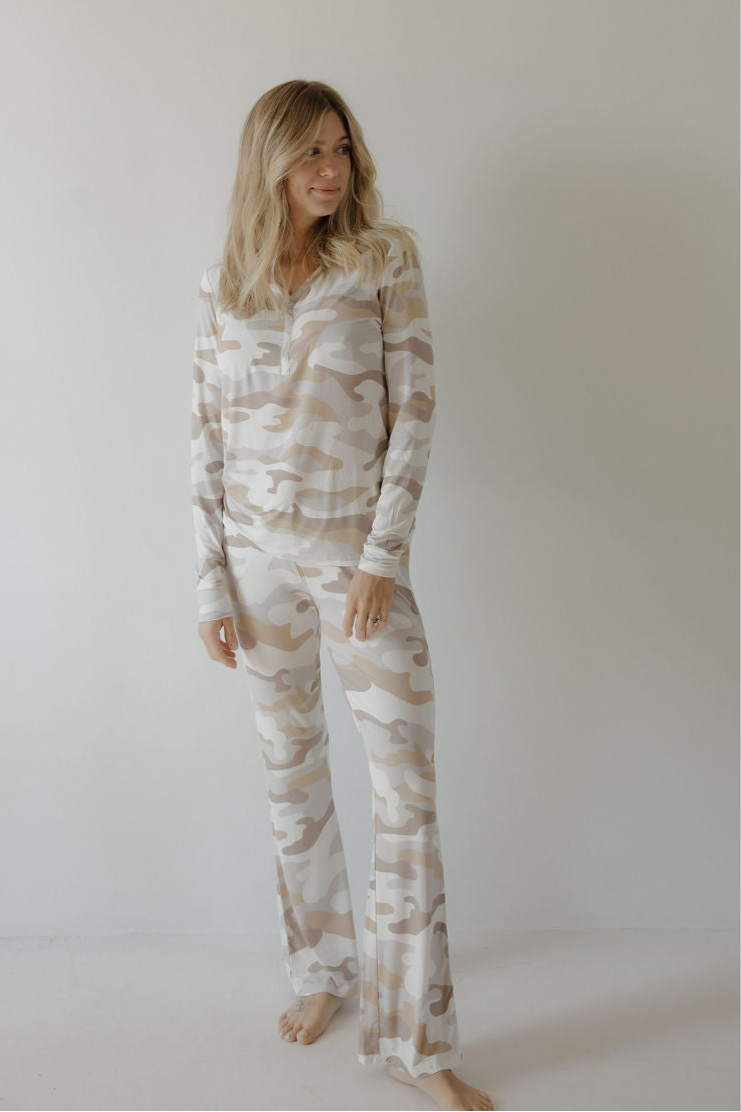 A woman with light brown hair wears forever french baby’s Women's Bamboo Pajama | Camo 2.0—long-sleeve, beige and white camo print. She stands barefoot against a white wall, softly smiling. The breathable bamboo fabric ensures all-night comfort.