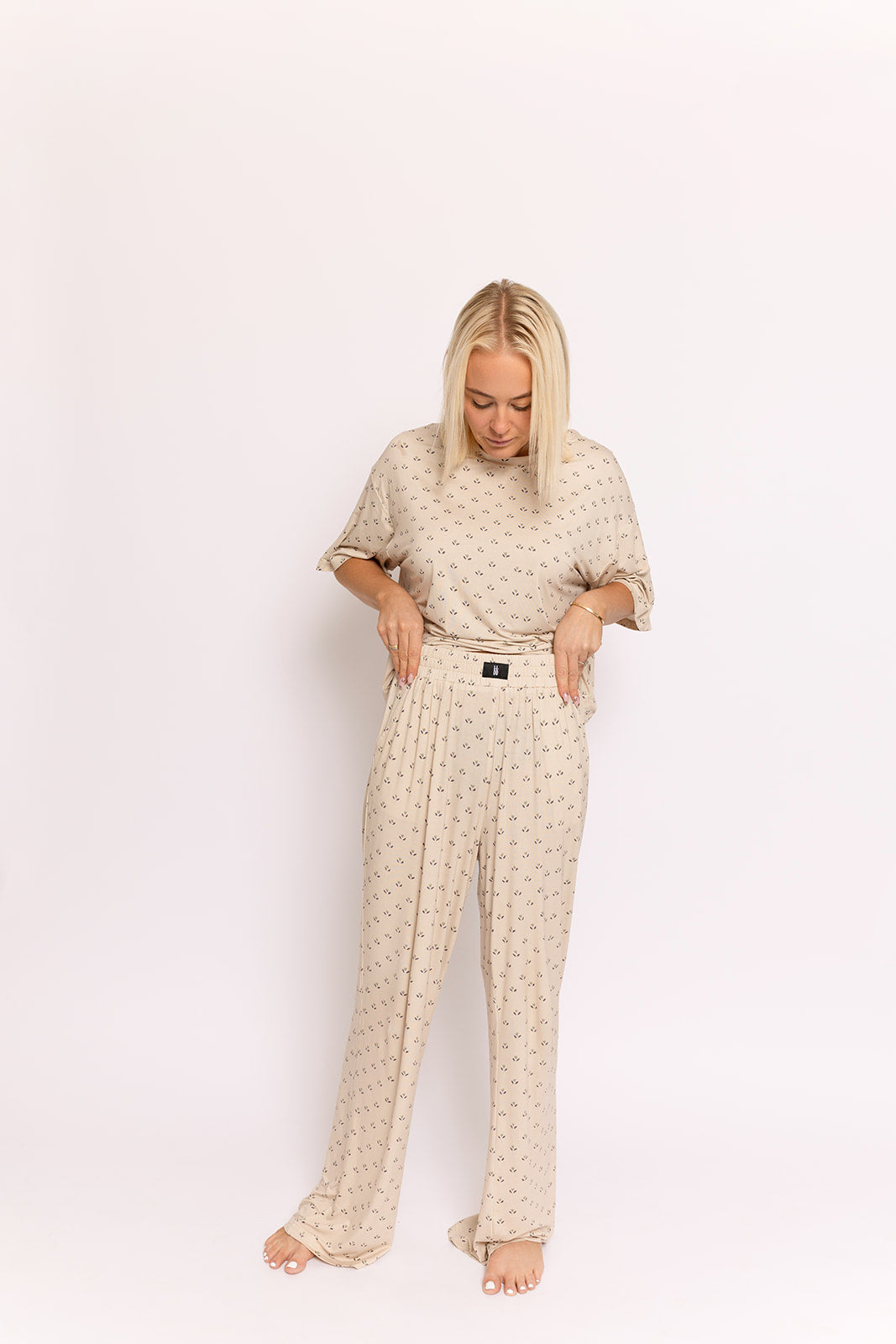 A blonde woman stands barefoot on a white background, looking down while wearing the forever french baby Women's Boxy Tee Lounge Set | Flora with matching beige patterned bamboo fabric, loose-fit pants, and a visible black waistband tag.