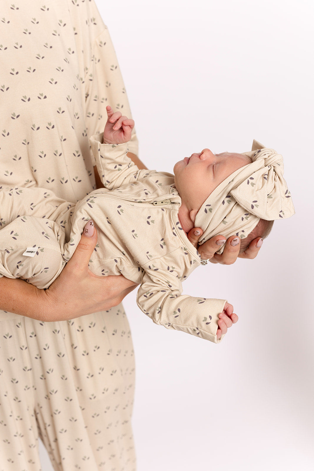 An adult holds a newborn dressed in forever french baby's Bamboo Zip Pajamas | Flora—a beige, leaf-patterned, breathable outfit with a matching headband—offering cozy comfort against a white background.