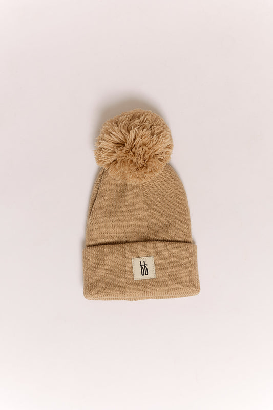 The ff Knit Pom Top Beanie | Taupe by forever french baby is a beige knit beanie with a large pom-pom, folded brim, and a small “bb” logo patch, shown on a plain, light background.