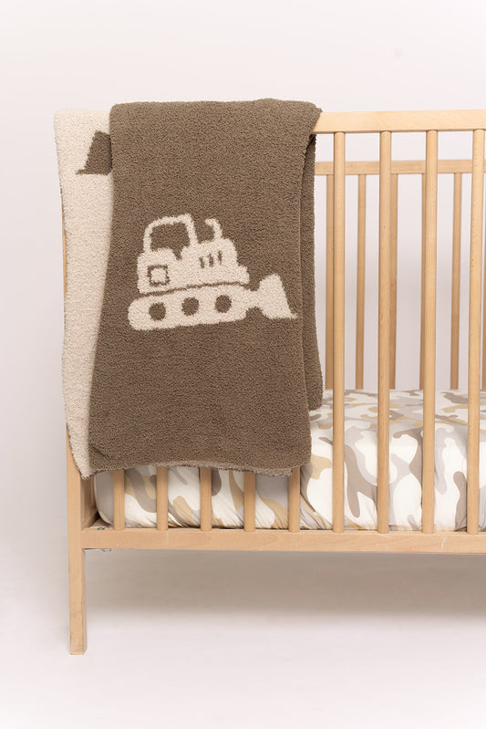 A wooden crib with a beige and white camo-patterned mattress is complemented by the forever french baby Plush Blanket | Bulldozer, a soft microfiber blanket with a bulldozer design draped over the side for comfort and style.