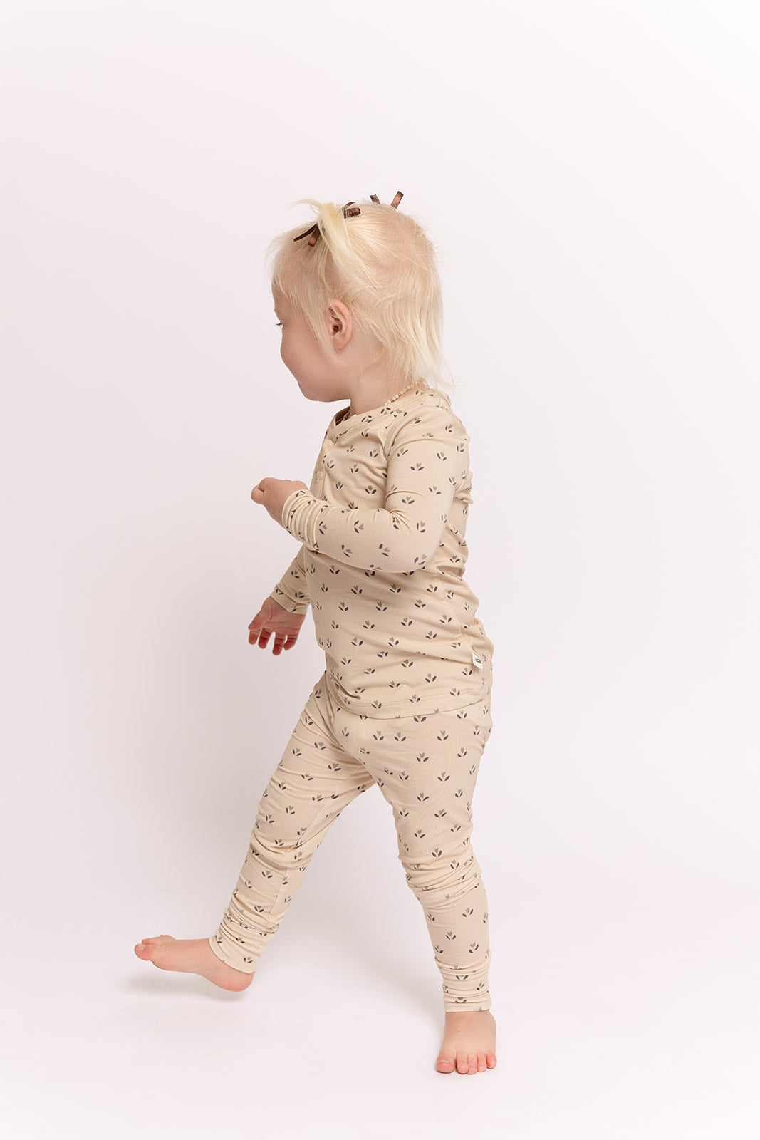A young child with light blonde hair wears the forever french baby Bamboo Two Piece Pajamas | Flora, a comfy beige set with black patterns, standing barefoot against a plain white background and looking to the side.