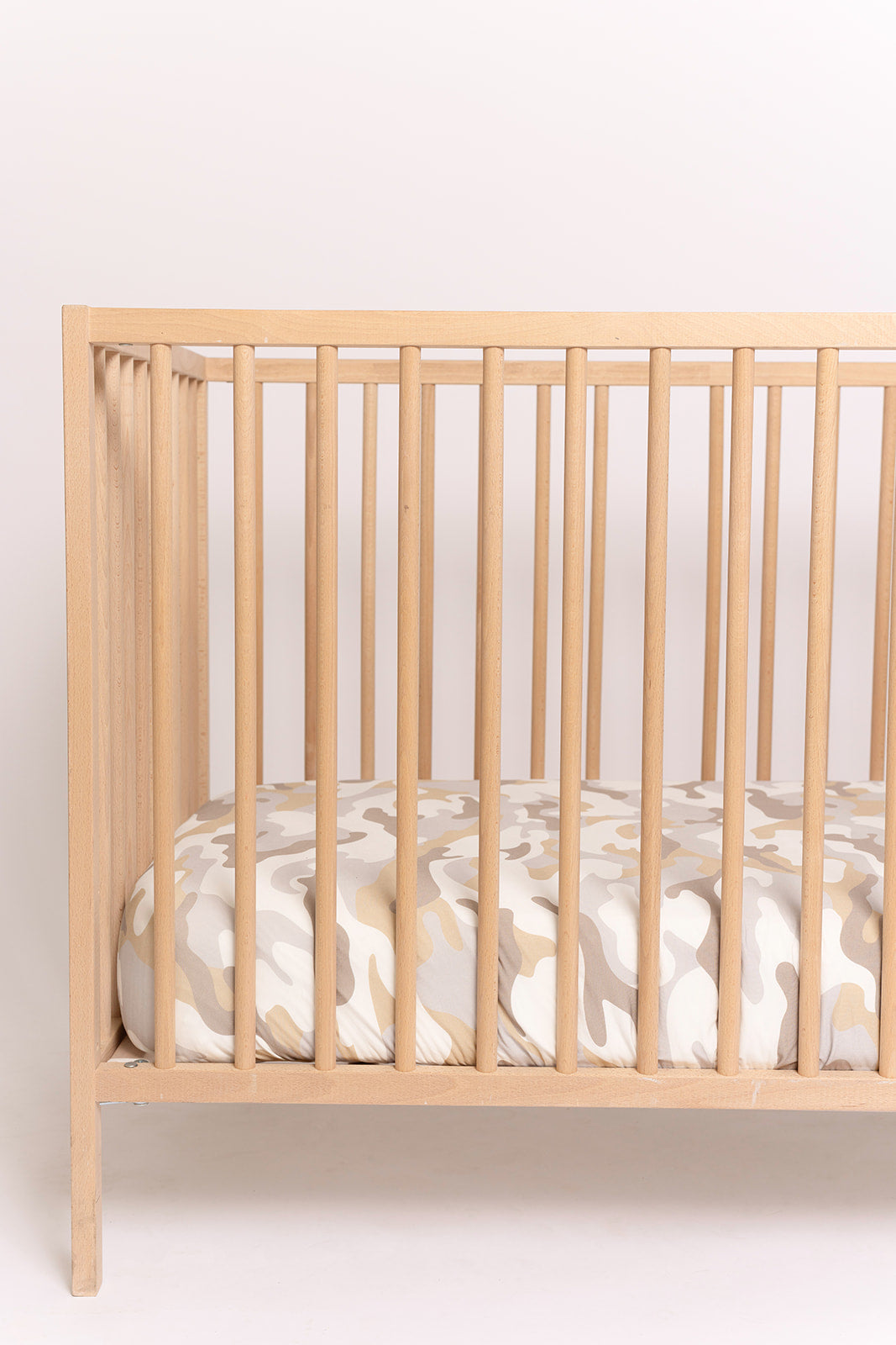 A light wooden baby crib with vertical slats holds a mattress covered in the forever french baby Bamboo Crib Sheet | Camo 2.0, featuring a beige and white camouflage pattern, all set against a plain white background.