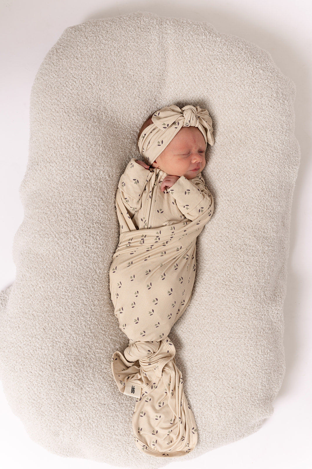 A newborn sleeps peacefully, swaddled in the forever french baby Bamboo Swaddle | Flora with a matching headband, resting on a soft, light cushion against a white background.