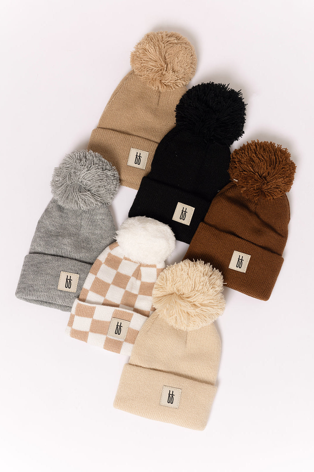 Six forever french baby ff Knit Pom Top Beanies in beige, black, gray, brown, cream, and beige-checkered patterns are displayed on a white background. Each beanie features a classic ribbed design, playful pom-pom top, and logo tag on the cuff.
