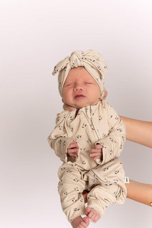 A newborn in forever french baby Bamboo Zip Pajamas | Flora—a beige, leaf-patterned sleepwear set with matching headwrap—is gently held upright by an adult against a plain, light background.