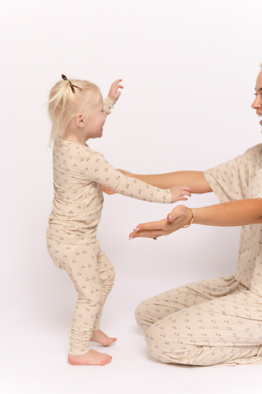 A toddler in forever french baby's Bamboo Two Piece Pajamas | Flora walks toward an adult in matching pajamas, both wearing the beige set with small patterns, against a plain white background, arms outstretched to greet each other.