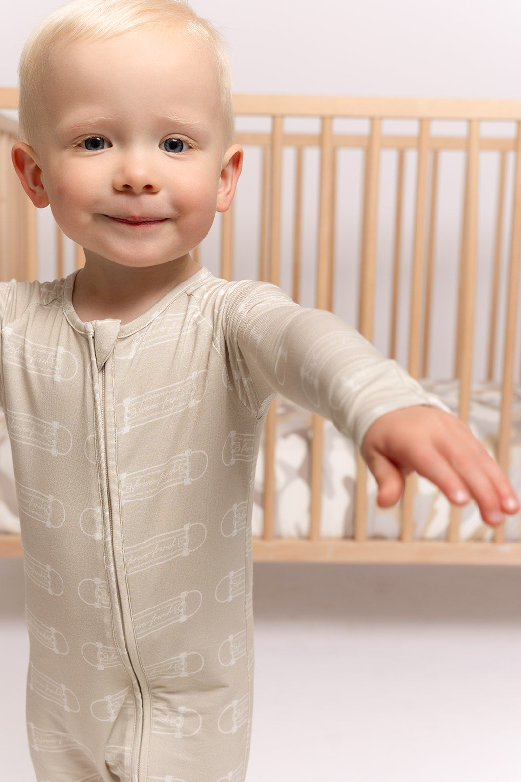 A smiling toddler with light blond hair wears forever french baby Bamboo Zip Pajamas | Sk8er Boy, standing with arms outstretched in front of a wooden crib in a softly lit, cozy room.