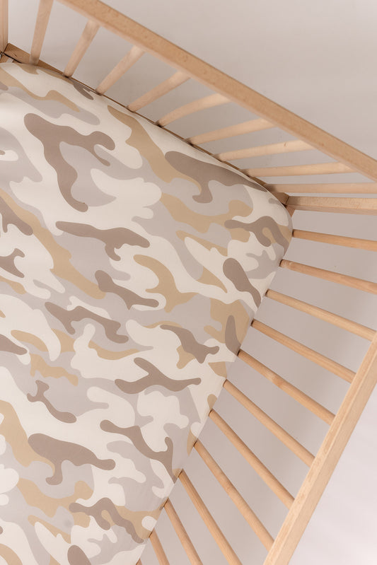 A wooden crib holds a baby mattress covered with the forever french baby Bamboo Crib Sheet in Camo 2.0, featuring a beige, tan, and cream camouflage pattern. The angled overhead shot highlights the stylish nursery bedding.