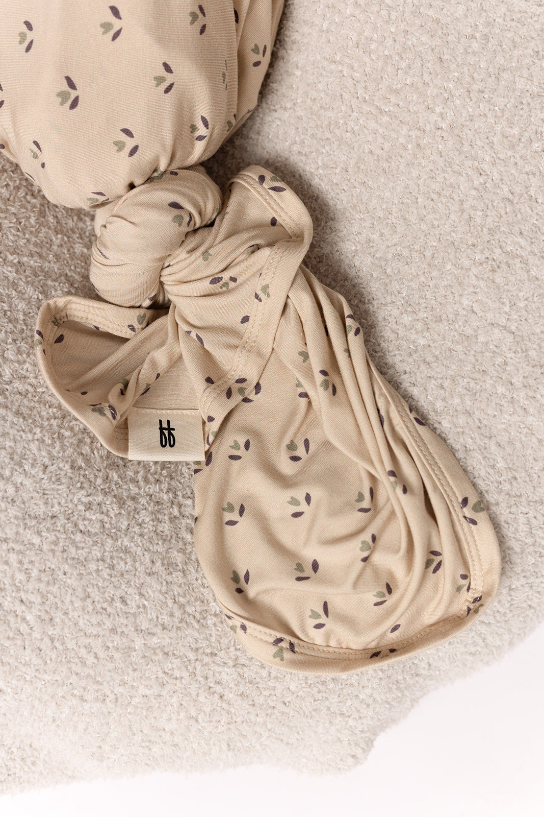 A close-up of the Bamboo Swaddle | Flora by forever french baby, featuring a beige fabric with small purple and green leaf patterns, tied in a knot on a light textured surface. A white tag with black “bb” lettering is visible.