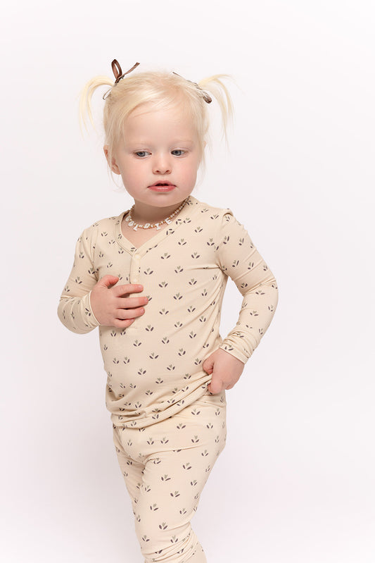 A toddler with blonde pigtails models the forever french baby Bamboo Two Piece Pajamas | Flora, standing against a plain white background, gazing slightly to the side with one hand on their chest and the other on their hip.