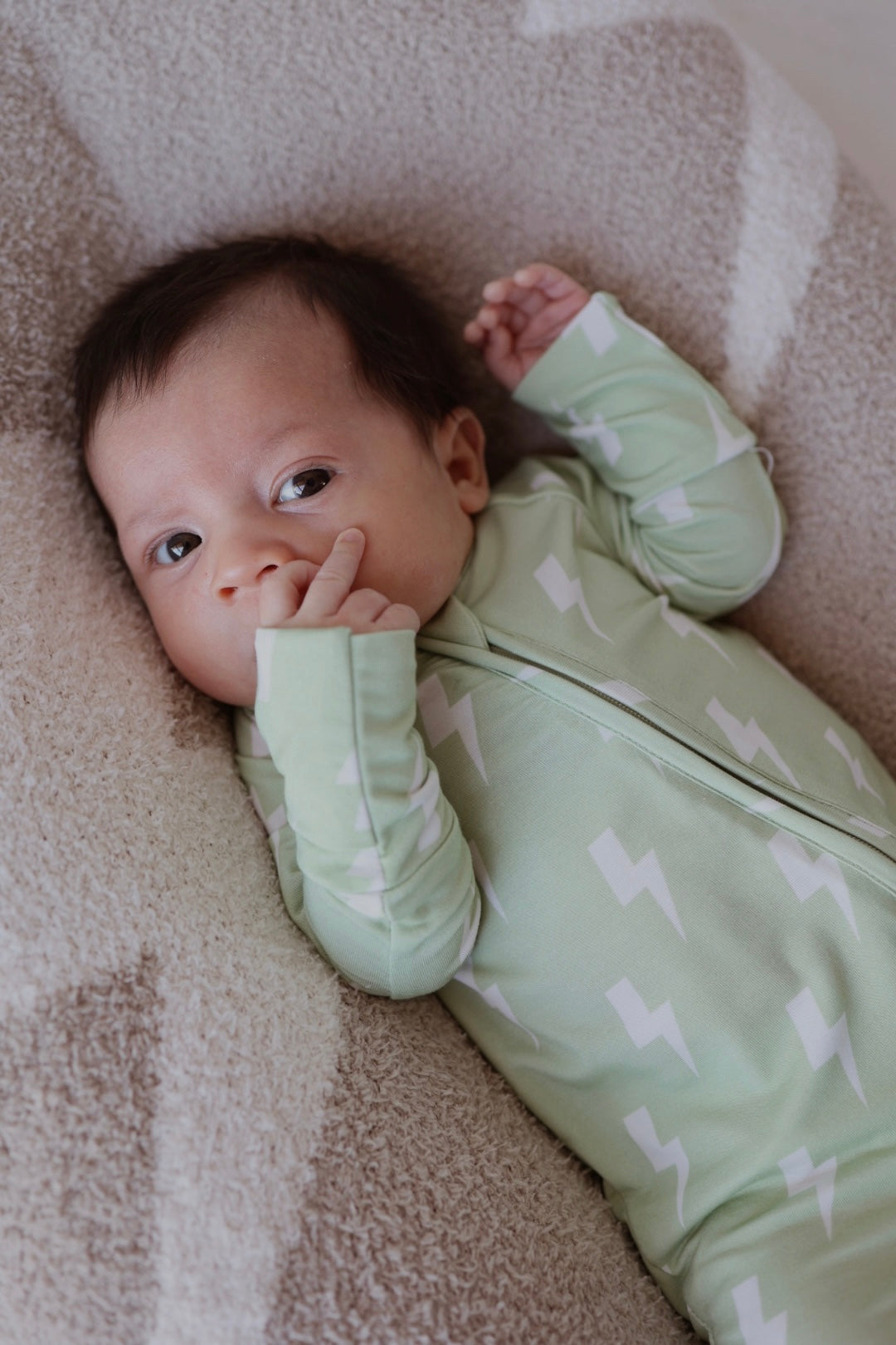 A dark-haired baby wearing forever french baby Bamboo Zip Pajamas | Flash—a light green onesie with white lightning bolts—lies on a soft, textured blanket, sucking their thumb.