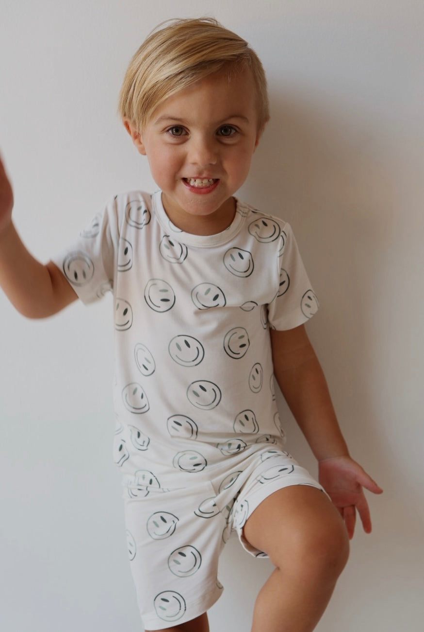 A young child with blonde hair smiles against a plain white background, wearing the forever french baby Bamboo Short Set | Green Smiles—breathable bamboo pajamas decorated with a playful smiley face pattern.