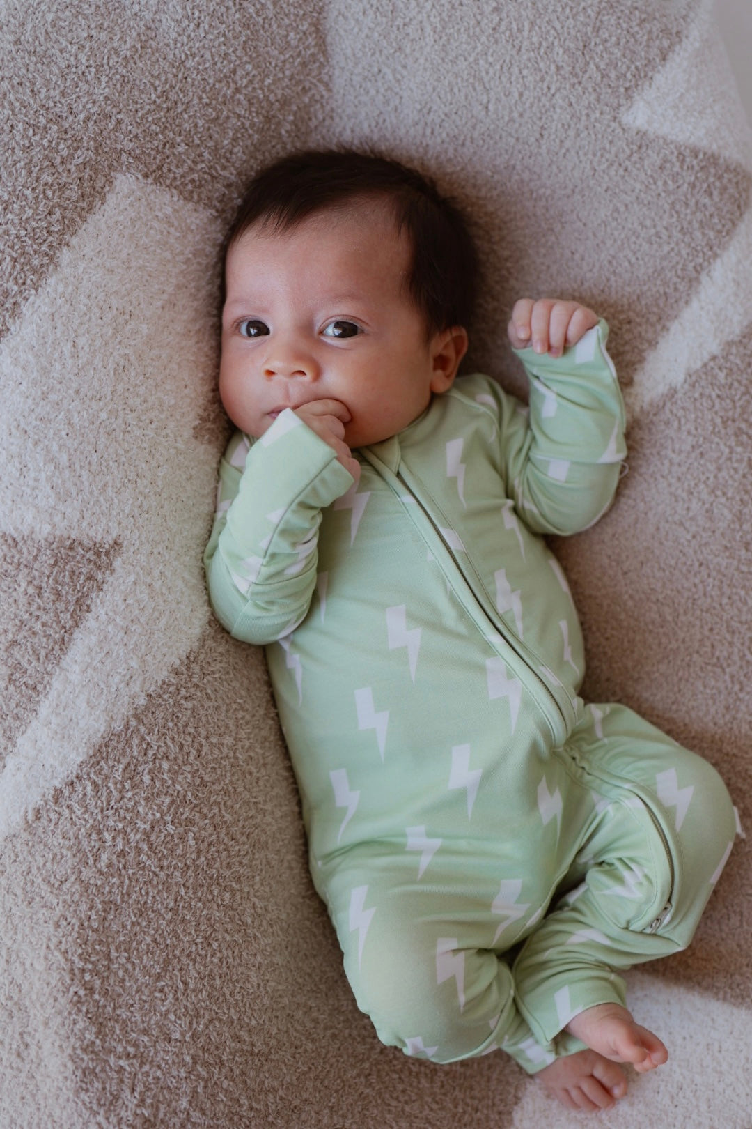 A baby in forever french baby's Bamboo Zip Pajamas | Flash, a light green onesie with white lightning bolts, lies on a beige star-patterned blanket and looks at the camera with one hand near their mouth.