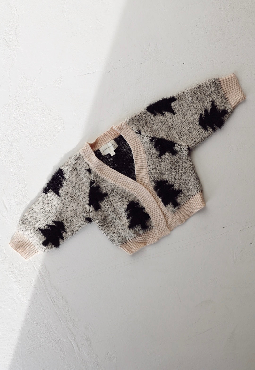 A children's "Cozy Cardigan | Silent Night" by forever french baby is displayed on a white surface. The cardigan features a light beige base with black abstract patterns, V-neck, and ribbed cuffs. Shadows create a diagonal line across the image, adding depth to this elegant and soft piece.